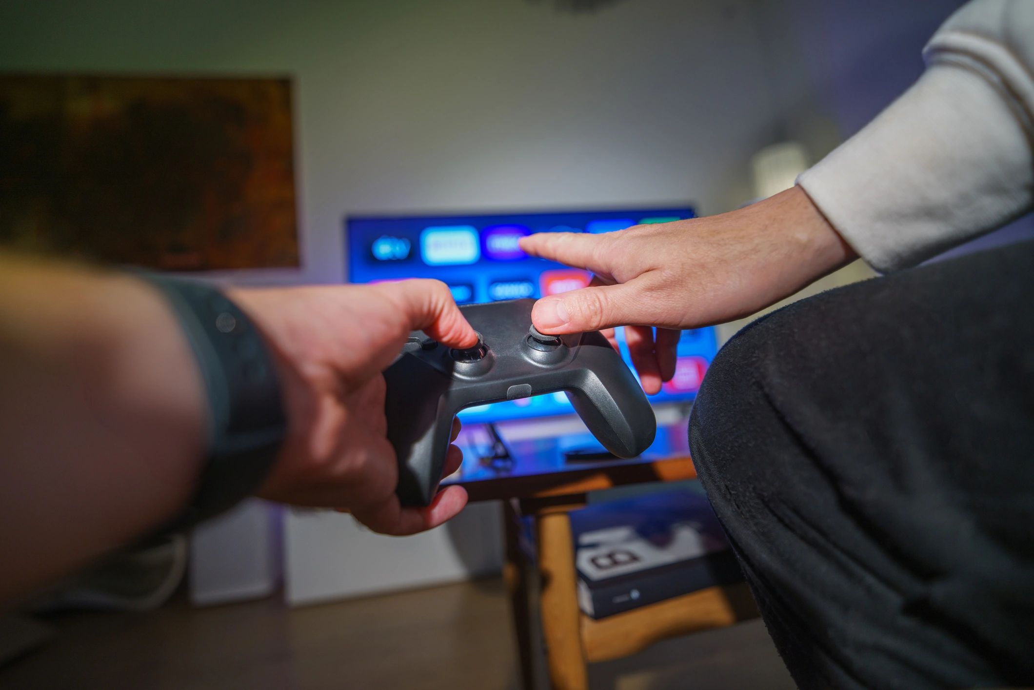 Hands reaching for a video game controller in front of a TV screen.